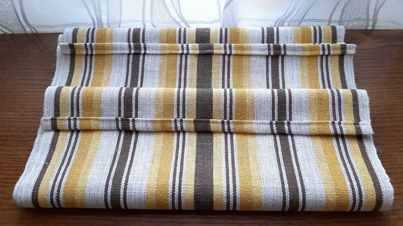 May include: A striped table runner with brown and yellow stripes on a white background. The runner is folded in thirds and is lying on a wooden surface.