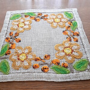 May include: A square, beige linen placemat with a floral embroidery design. The design features yellow flowers with orange centers and green leaves, all arranged in a circular pattern around the center of the placemat.