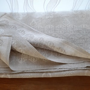 May include: A folded linen tablecloth with a white damask pattern. The tablecloth is a light beige color and has a subtle sheen.