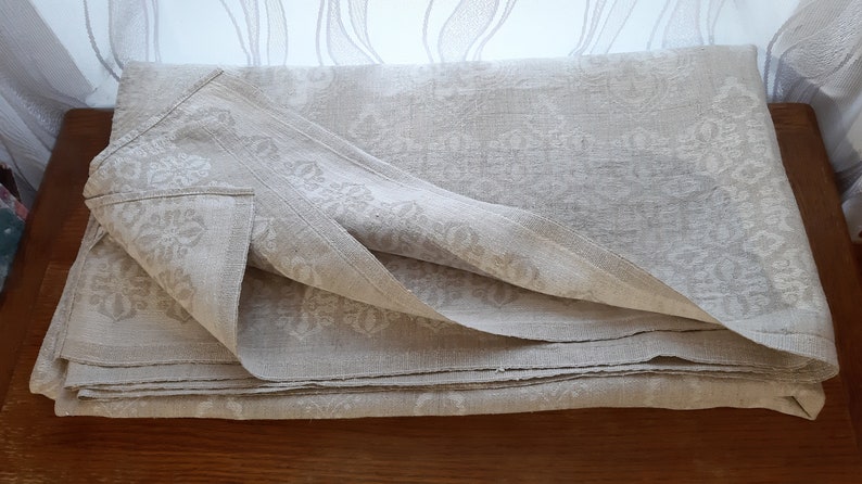May include: A folded linen tablecloth with a white damask pattern. The tablecloth is a light beige color and has a subtle sheen.