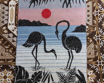 Op de afbeelding: Geweven wandkleed met een zonsondergangscène met twee zwarte flamingo's in een kustlandschap. De achtergrond omvat een rode zon, een roze en blauwe lucht en zwarte silhouetten van eilanden en palmbladeren. Het wandtapijt is omzoomd door houten deuvels.