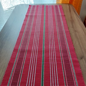 May include: Red and green striped table runner with a woven texture. The runner is made of a fabric material and has a rectangular shape.