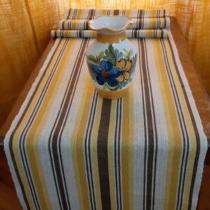 May include: A yellow, white, and brown striped table runner with a floral patterned ceramic vase in the center.