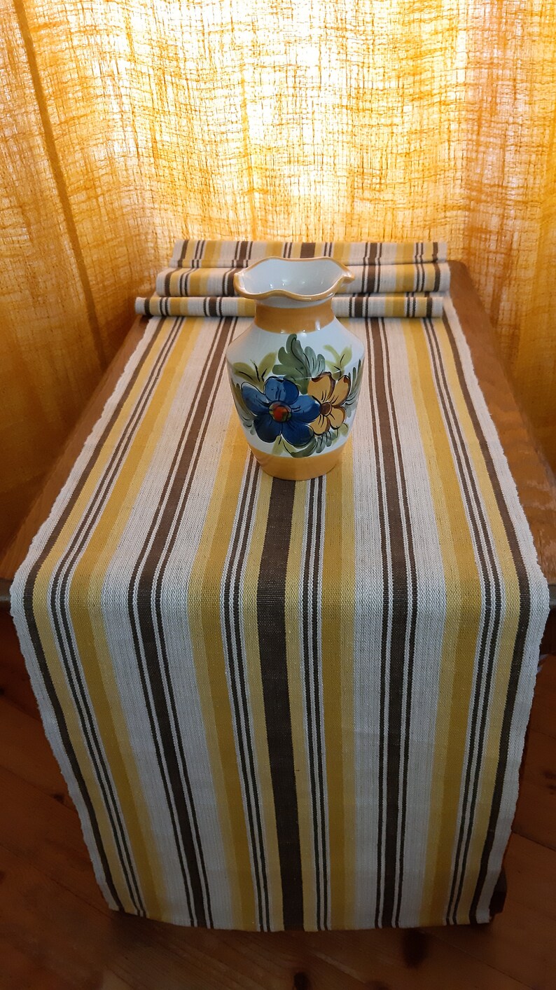 May include: A yellow, white, and brown striped table runner with a floral patterned ceramic vase in the center.