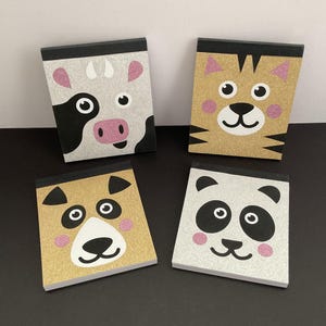 May include: Four square notepads featuring animal faces: a cow, tiger, bear, and panda. The cow and panda have silver glitter backgrounds, while the tiger and bear have gold glitter. Each notepad has a black border and pink accents.
