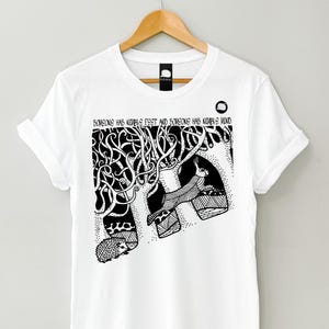 Può includere: Maglietta bianca con un'illustrazione in bianco e nero di un albero con un coniglio e un riccio. Il testo "Someone has nimble feet and someone has nimble mind" è stampato sulla maglietta.