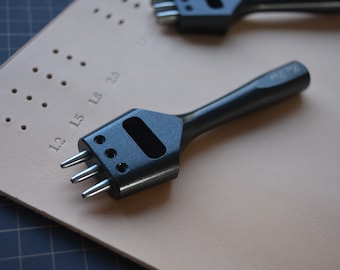 Puede incluir: Una herramienta de punzonado de cuero negra con tres puntas, utilizada para hacer agujeros en el cuero. La herramienta está sobre un trozo de cuero marrón con marcas numeradas.