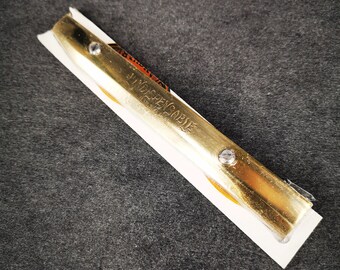 Op de afbeelding: Een gouden metalen scheermesje met de woorden "Indestructible" gegraveerd op het mesje. Het mesje is bevestigd aan een wit kunststof handvat.
