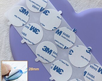 Könnte beinhalten: Nahaufnahme einer Packung 3M doppelseitiges Gewebband. Das Band ist weiß mit blauer Beschriftung und einem 3M-Logo. Das Band hat einen Durchmesser von 28 mm.