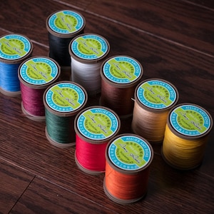 Pode incluir: Dez rolos de fio de várias cores, incluindo vermelho, verde, laranja, amarelo e marrom. Cada rolo tem um rótulo que diz "Atelier Amy Roke" e "Polyester" em inglês e japonês. Os rolos estão dispostos em círculo sobre uma superfície de madeira.