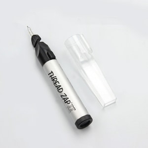 Może przedstawiać: Srebrno-czarny palnik do nici Thread Zap II z przezroczystą plastikową nasadką. Narzędzie ma spiczastą końcówkę, a na srebrnym korpusie wydrukowano napis "THREAD ZAP II".