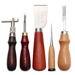 Op de afbeelding: Een verzameling leerbewerkingsgereedschappen met houten handvatten en metalen bladen. De gereedschappen omvatten een draaimes, een groover, een afsteekmes met Japanse tekst, een priem en een stikgroover. De handvatten zijn in verschillende bruintinten.