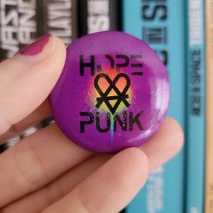 Könnte beinhalten: Lilafarbener Knopf mit den Worten "HOPE PUNK" in schwarzer Schrift. Ein Regenbogenherz mit einem schwarzen X darüber befindet sich in der Mitte des Knopfes.