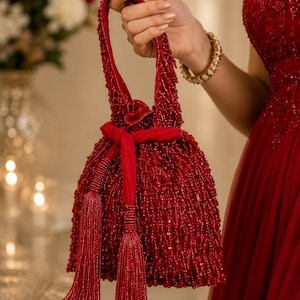 May include: A deep red drawstring handbag covered in dense, shimmering beads. The bag features a matching red fabric tie closure, a single beaded top handle, and two long, beaded tassel accents hanging from the front, creating a textured look.