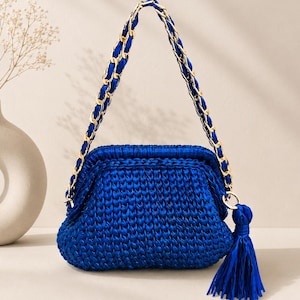 Peut inclure: Un sac à main en crochet bleu roi vibrant doté d'une fermeture à cadre structurée et d'une bandoulière en chaîne dorée entrelacée de ruban bleu assorti. Un long pompon bleu épais est suspendu à l'anneau latéral, ajoutant un élément décoratif.