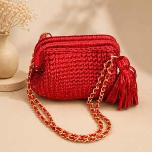 May include: A vibrant red woven clutch bag featuring a structured frame, a gold-tone chain shoulder strap interwoven with matching red cord, and a large, decorative red tassel attached to the side. The bag has a textured, braided appearance.