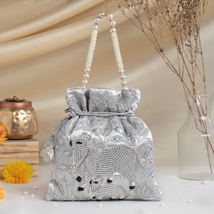 Puede incluir: Un bolso tipo saco plateado con cierre de cordón, decorado con abalorios que representan una vaca y un ternero, junto con bordados florales. Incluye un asa de cuentas blancas tipo perla y una borla plateada a juego en el cordón lateral.