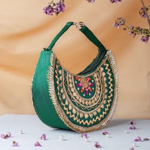 Puede incluir: Un bolso de mano verde esmeralda en forma de media luna con una sola asa superior y cierre de cremallera. El exterior está decorado con ribetes metálicos dorados, detalles reflectantes tipo espejo y un motivo floral central en hilo.