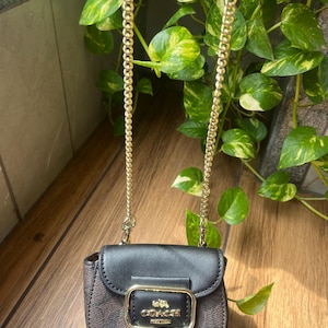 Op de afbeelding: Een kleine crossbodytas van het merk Coach met een donkerbruine body met monogrampatroon en een zwarte leren klep. De tas is voorzien van een goudkleurig metalen gespdetail met het Coach-logo en een lange goudkleurige kettingriem.