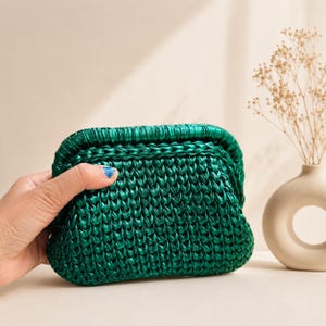 May include: A small, rectangular clutch bag crafted from metallic emerald green yarn in a dense, textured crochet stitch. The bag features a structured frame closure at the top, with a consistent, shiny finish throughout the woven material.