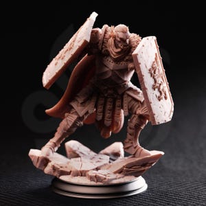 May include: A detailed figurine of a warrior in a dynamic pose, holding two shields. The figure is clad in armor and a cape, standing on a rocky base. The shields feature embossed designs, and the overall color is a muted brown.
