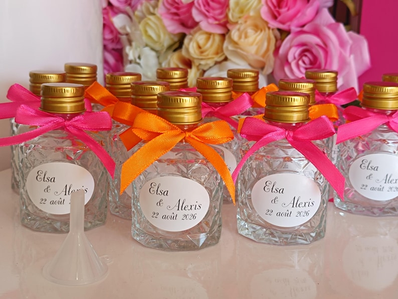 May include: Small, clear glass bottles with gold lids, decorated with pink and orange ribbons. Each bottle has a label with the names "Elsa & Alexis" and the date "22 ao&ucirc;t 2026". A white funnel is in the foreground, with a backdrop of pink and yellow roses.