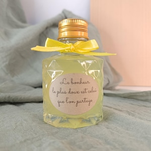 May include: A small, clear glass bottle filled with a yellow liquid, topped with a gold cap and a yellow ribbon. The bottle has a diamond-patterned texture and a label with French text.
