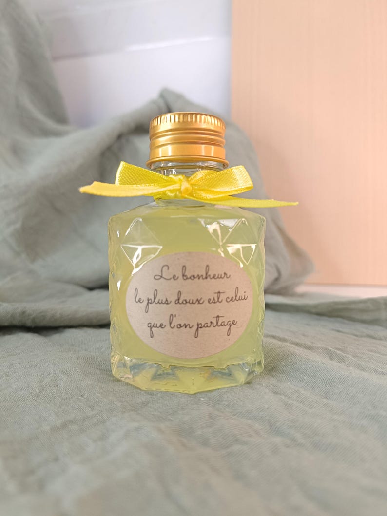 May include: A small, clear glass bottle filled with a yellow liquid, topped with a gold cap and a yellow ribbon. The bottle has a diamond-patterned texture and a label with French text.