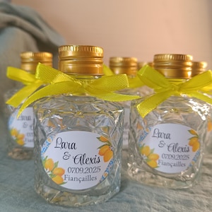 May include: Small, clear glass bottles with gold-coloured caps and yellow ribbons. Each bottle has a label with the names "Lara & Alexis", the date "07.09.2025", and the word "Fiancailles" with a lemon illustration. The bottles are arranged on a light green surface.