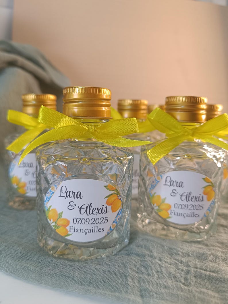 May include: Small, clear glass bottles with gold-coloured caps and yellow ribbons. Each bottle has a label with the names "Lara & Alexis", the date "07.09.2025", and the word "Fiancailles" with a lemon illustration. The bottles are arranged on a light green surface.