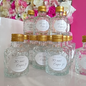 May include: A display of small, clear glass bottles with gold lids. Each bottle features a diamond pattern and a white label with the text "30 ans Capu" and the date "13 November 2021".