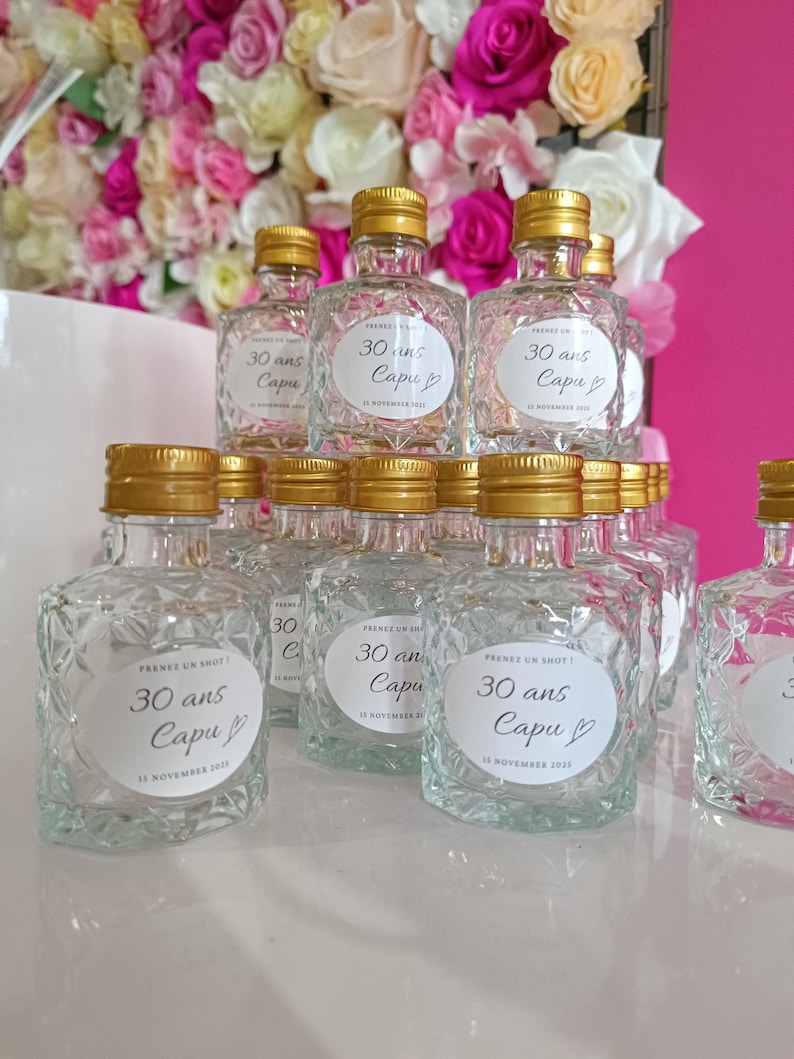May include: A display of small, clear glass bottles with gold lids. Each bottle features a diamond pattern and a white label with the text "30 ans Capu" and the date "13 November 2021".