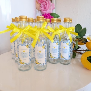 May include: A collection of small, clear glass bottles with gold screw caps, each tied with a bright yellow satin ribbon. The bottles feature rectangular labels with blue and white tile patterns, yellow lemon illustrations, and the text Olive & Jake.