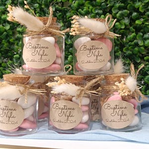 Peut inclure: Petits bocaux en verre remplis de bonbons roses et blancs, ornés de bouchons en liège, de ficelle et de fleurs séchées. Chaque bocal porte une étiquette avec "Baptême Nyla 12 octobre 2024". Les bocaux sont disposés en cascade.
