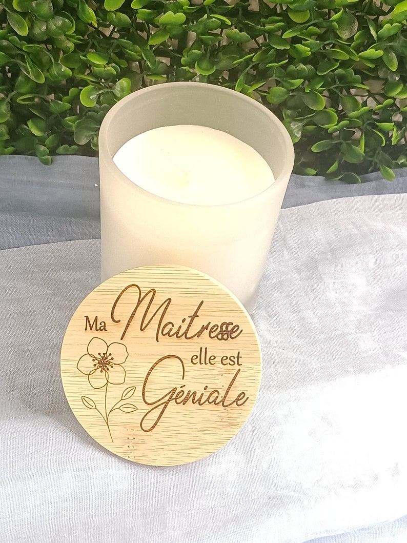 May include: A white frosted glass candle jar with a wooden lid. The lid has a floral design and the text "Ma Maitresse elle est Geniale".