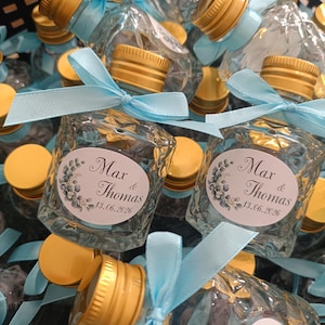 May include: Small, clear glass bottles with gold-coloured caps and light blue ribbons. Each bottle has a label with the names "Max & Thomas" and the date "13.06.2026". The bottles are arranged in a dark container.