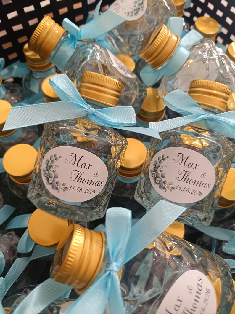 May include: Small, clear glass bottles with gold-coloured caps and light blue ribbons. Each bottle has a label with the names "Max & Thomas" and the date "13.06.2026". The bottles are arranged in a dark container.