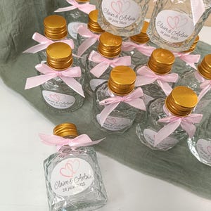 May include: Small, clear glass bottles with gold-coloured caps and pink ribbons. Each bottle has a label with the names "Claire & Arlain" and the date "28 juin 2025". The bottles are arranged on a light green fabric.