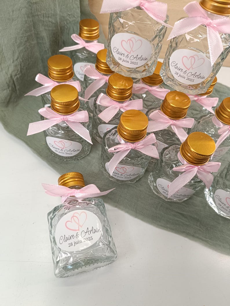 May include: Small, clear glass bottles with gold-coloured caps and pink ribbons. Each bottle has a label with the names "Claire & Arlain" and the date "28 juin 2025". The bottles are arranged on a light green fabric.