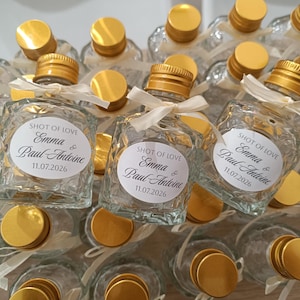 May include: Small, clear glass bottles with gold lids and white ribbon ties. Each bottle features a white label with the text "SHOT OF LOVE", the names "Emma & Paul Antoine", and the date "11.07.2026".
