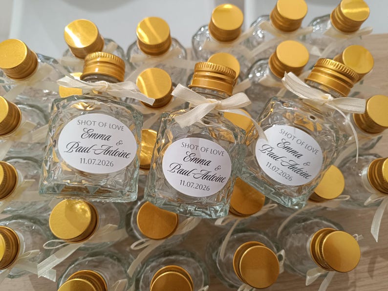 May include: Small, clear glass bottles with gold lids and white ribbon ties. Each bottle features a white label with the text "SHOT OF LOVE", the names "Emma & Paul Antoine", and the date "11.07.2026".