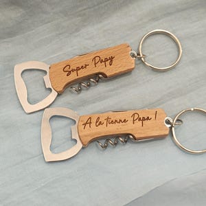Op de afbeelding: Twee houten flessenopeners met sleutelhangers. De bovenste flesopener heeft de tekst "Super Papy" gegraveerd. De onderste flesopener heeft de tekst "A la tienne Papa!" gegraveerd.
