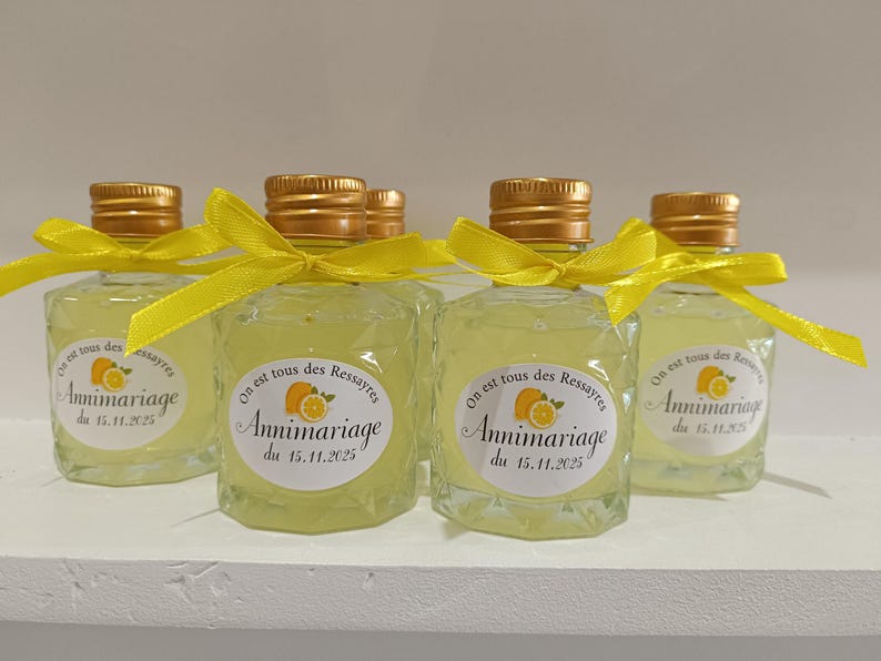 May include: Small, clear glass bottles filled with a yellow liquid, each topped with a gold cap and tied with a yellow ribbon. The bottles have a label with text and a citrus fruit illustration. The bottles are arranged on a white surface.