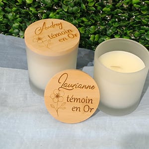 May include: Two white candles with wooden lids. The lids are engraved with the text "Audrey t&eacute;moin en Or" and "Laurianne t&eacute;moin en Or".