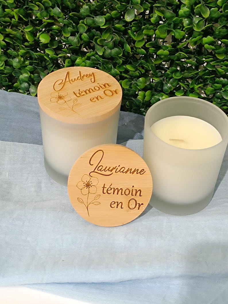 May include: Two white candles with wooden lids. The lids are engraved with the text "Audrey t&eacute;moin en Or" and "Laurianne t&eacute;moin en Or".
