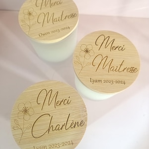 May include: Two round wooden candle lids with engraved text. The lids are light brown with a floral design and the text "Merci Maitresse" and "Merci Charlene" with the year "2023-2024".