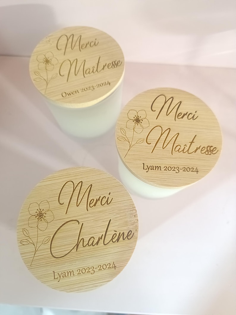 May include: Two round wooden candle lids with engraved text. The lids are light brown with a floral design and the text "Merci Maitresse" and "Merci Charlene" with the year "2023-2024".