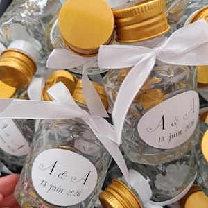 May include: Small glass bottles with gold-coloured caps, tied with white ribbons. Each bottle has a white label with the text "A & A 13 juin 2026". The bottles are arranged in a black container.