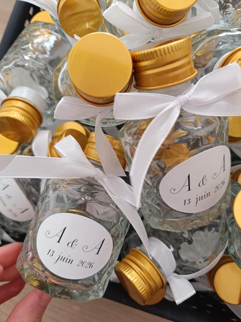 May include: Small glass bottles with gold-coloured caps, tied with white ribbons. Each bottle has a white label with the text "A & A 13 juin 2026". The bottles are arranged in a black container.