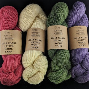 May include: Four skeins of yarn in different colors: red, cream, green, and purple. Each skein has a brown label with the text "The Ornery Shepherd Gulf Coast Native Wool Yarn".
